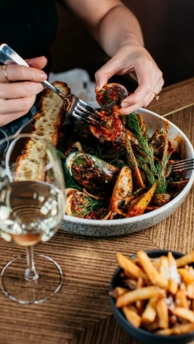 A lady eating mussels from a bowl, with a glass of wine and side bowl of chips