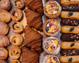 Looking down at a table of delicious pastries in rows, chocolate and caramel eclairs, Portuguese tarts, croissants, scrolls and cruffins.