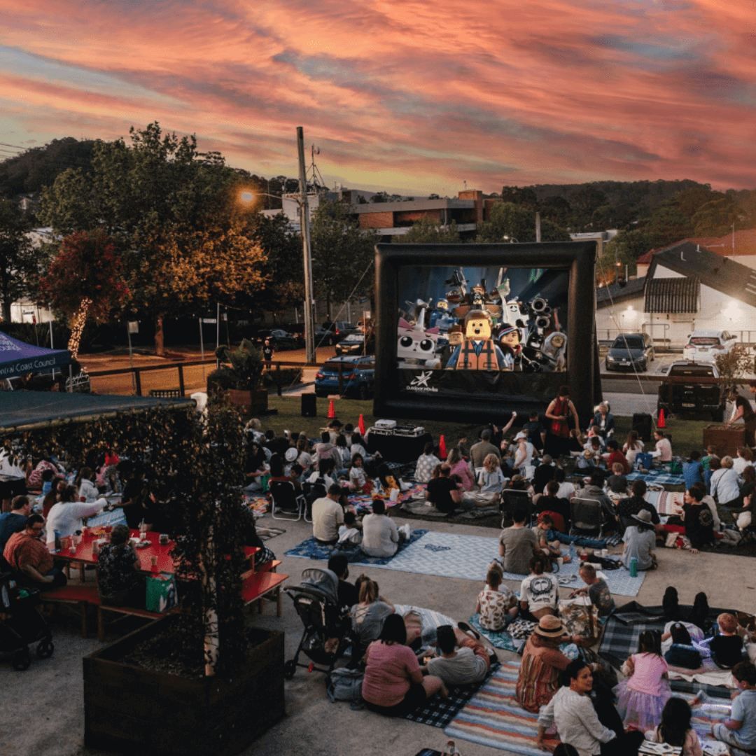 A crowd sits outside watching amovie on a large screen with a sunseet in the background